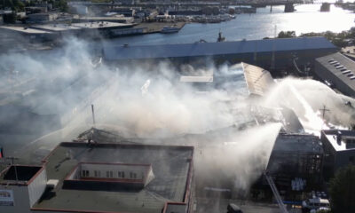 Aerial view of an industrial complex with thick white smoke and firefighters spraying water on a fire, with a harbor visible in the background