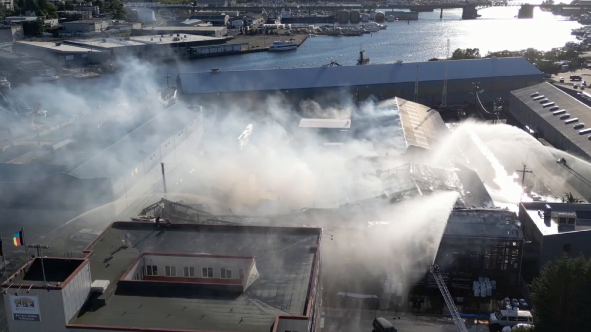 Aerial view of an industrial complex with thick white smoke and firefighters spraying water on a fire, with a harbor visible in the background