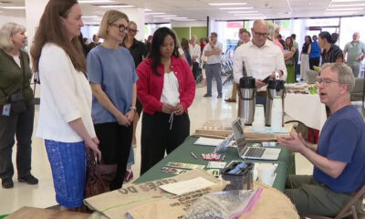 Instructor in a blue shirt explains at a table with laptop and coffee dispensers to a standing group in a bright room.