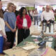 Instructor in a blue shirt explains at a table with laptop and coffee dispensers to a standing group in a bright room.