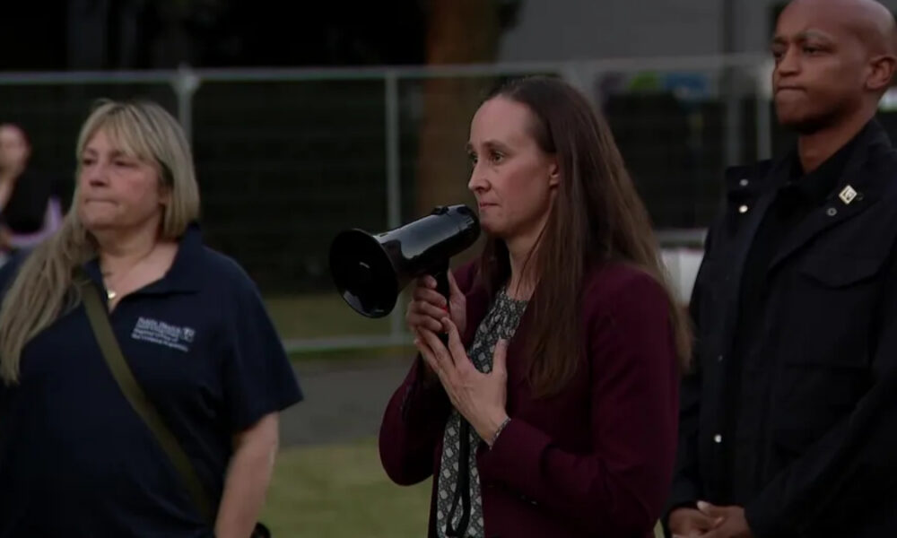Woman in a burgundy blazer speaks into a megaphone at an outdoor event, flanked by two people nearby.