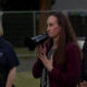 Woman in a burgundy blazer speaks into a megaphone at an outdoor event, flanked by two people nearby.