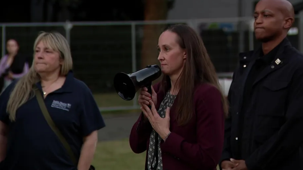 Woman in a burgundy blazer speaks into a megaphone at an outdoor event, flanked by two people nearby.