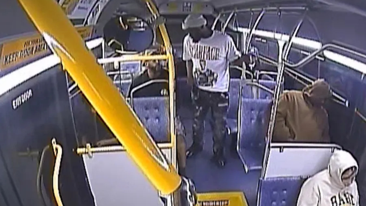 People standing and sitting inside a city bus with yellow handrails and blue seats, viewed from a security camera angle.