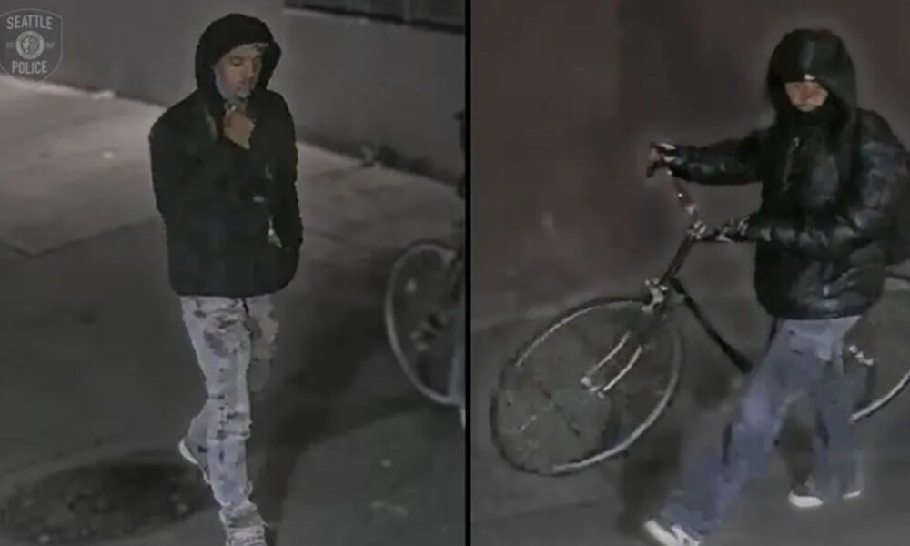 Surveillance image of a man in a black hoodie and jacket with light jeans, standing by a wall and a bicycle in the background (Seattle Police logo seen).