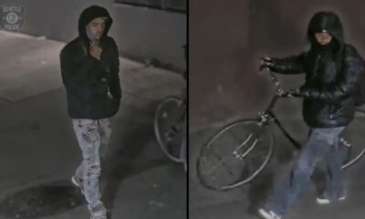 Surveillance image of a man in a black hoodie and jacket with light jeans, standing by a wall and a bicycle in the background (Seattle Police logo seen).