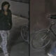Surveillance image of a man in a black hoodie and jacket with light jeans, standing by a wall and a bicycle in the background (Seattle Police logo seen).