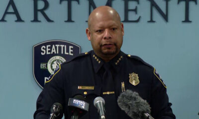 Police chief in uniform speaking at a press conference, Seattle Police badge visible behind him and multiple microphones in front of him.