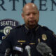 Police chief in uniform speaking at a press conference, Seattle Police badge visible behind him and multiple microphones in front of him.