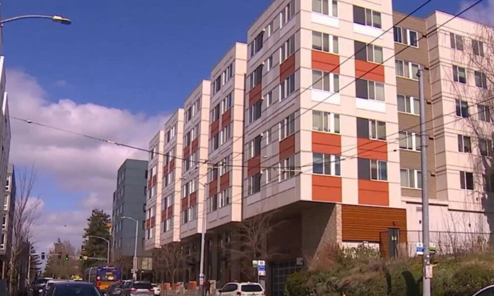 Five-story apartment building with white and red panels along a city street; cars parked below under a blue sky.