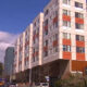 Five-story apartment building with white and red panels along a city street; cars parked below under a blue sky.