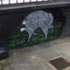 Graffiti of a gray boar walking on green ground painted on a dark brick wall in an alley, near a drain and trash bin.