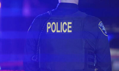 Back view of a police officer wearing a dark tactical vest with the word POLICE in yellow on the back, blue emergency lights visible in the surroundings.