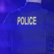 Back view of a police officer wearing a dark tactical vest with the word POLICE in yellow on the back, blue emergency lights visible in the surroundings.