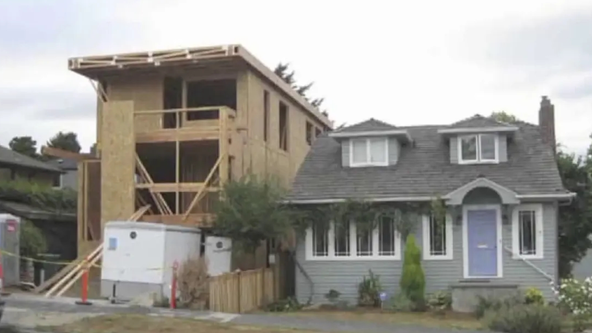 Wood-framed two-story house under construction beside a completed gray bungalow with a purple front door.