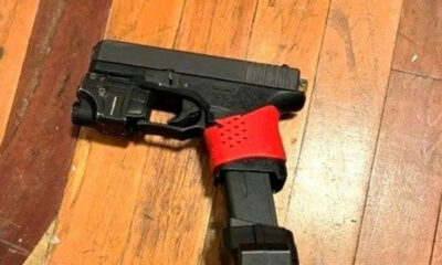 Black semi-automatic handgun on a wooden floor with a red perforated strap around the grip area holding the gun in place.