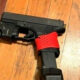 Black semi-automatic handgun on a wooden floor with a red perforated strap around the grip area holding the gun in place.
