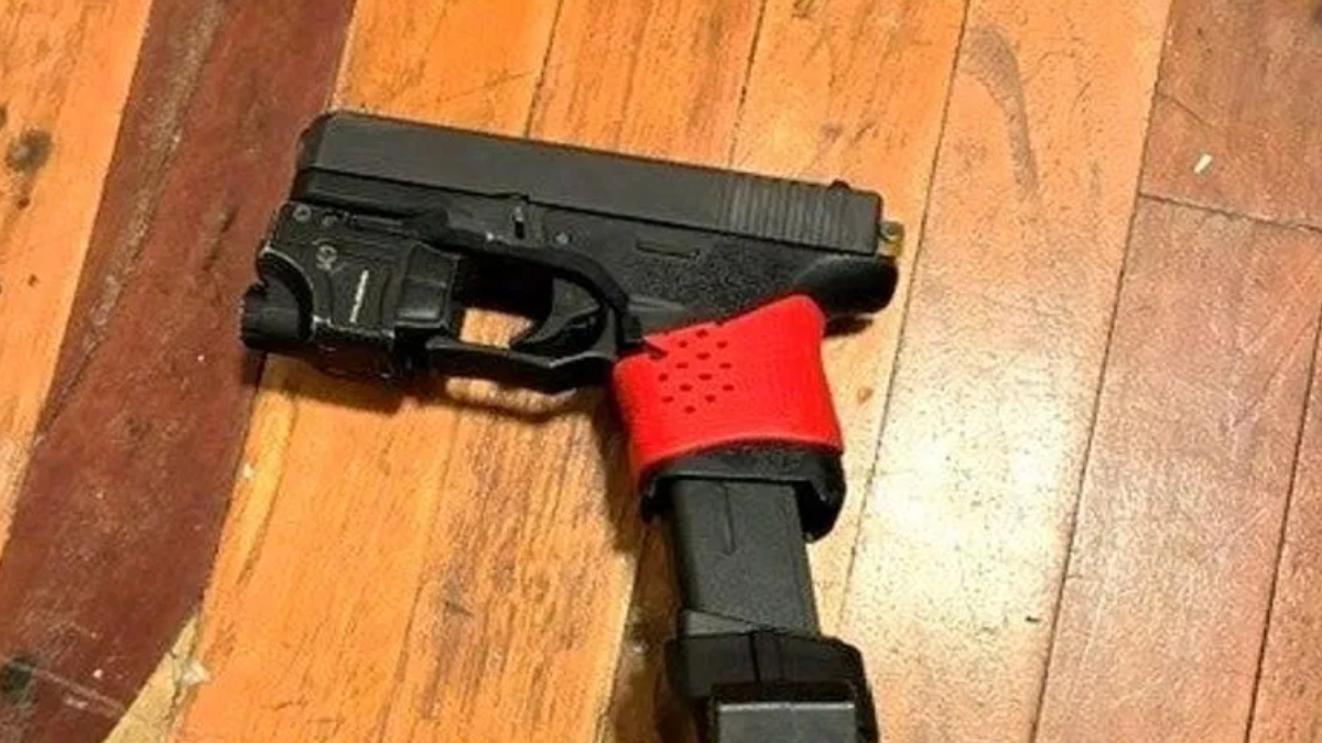 Black semi-automatic handgun on a wooden floor with a red perforated strap around the grip area holding the gun in place.