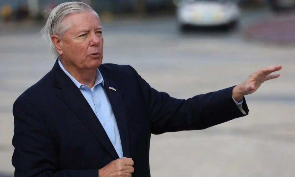 Older man in a navy blazer extends his right arm while speaking outdoors, background blurred.