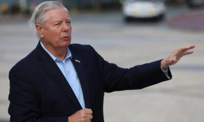 Older man in a navy blazer extends his right arm while speaking outdoors, background blurred.