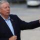 Older man in a navy blazer extends his right arm while speaking outdoors, background blurred.