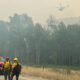 Firefighters in yellow gear walk near smoke-filled forest with a helicopter overhead and red trucks nearby on a dirt road.