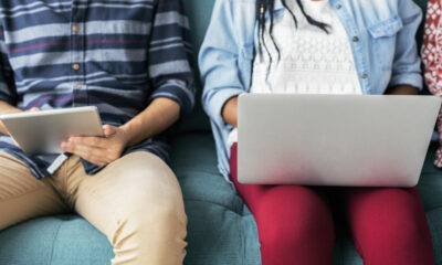 Two people sitting on a teal sofa using devices: one with a tablet, the other with a laptop on their laps.