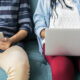 Two people sitting on a teal sofa using devices: one with a tablet, the other with a laptop on their laps.