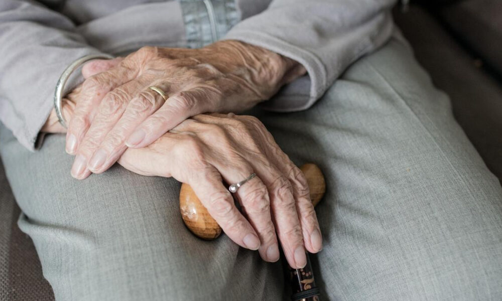 Close-up of an elderly person’s hands folded over a wooden cane, wearing rings, conveying aging and care.