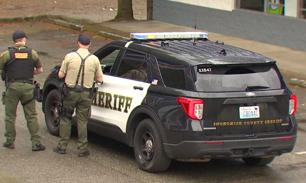 Two sheriff deputies stand beside a black-and-white sheriff patrol SUV with emergency lights in a parking lot.
