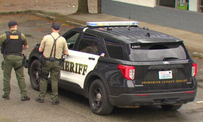 Two sheriff deputies stand beside a black-and-white sheriff patrol SUV with emergency lights in a parking lot.