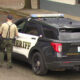 Two sheriff deputies stand beside a black-and-white sheriff patrol SUV with emergency lights in a parking lot.