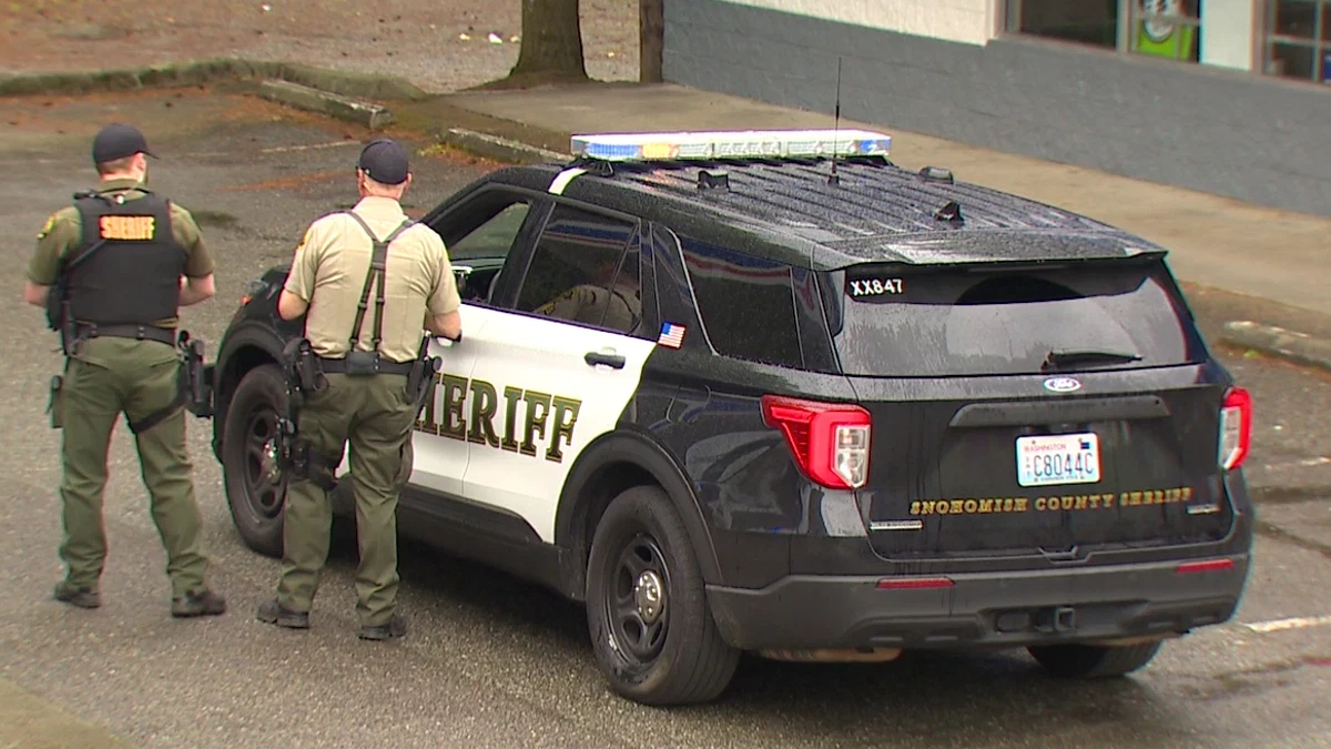 Two sheriff deputies stand beside a black-and-white sheriff patrol SUV with emergency lights in a parking lot.
