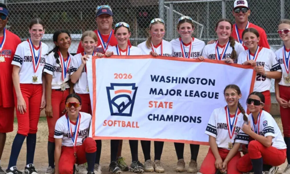Group of girls and coaches in red and white softball uniforms pose with a banner reading '2026 Washington Major League State Champions' and wearing medals behind a chain-link fence.