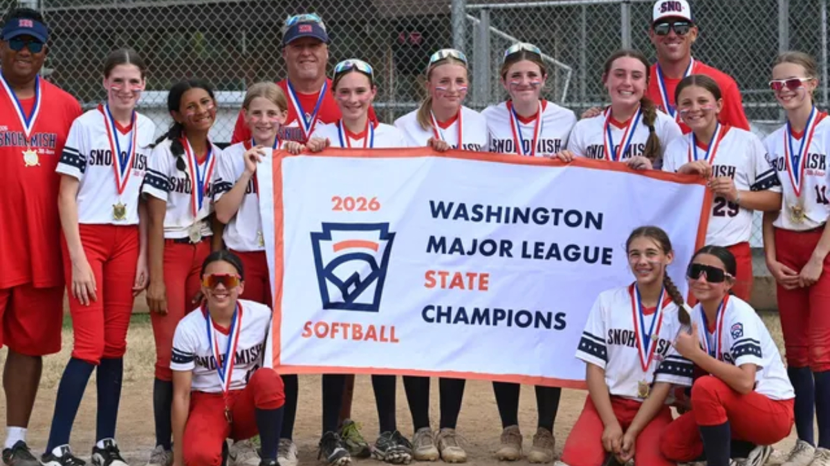 Group of girls and coaches in red and white softball uniforms pose with a banner reading '2026 Washington Major League State Champions' and wearing medals behind a chain-link fence.