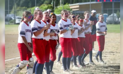 Girls softball team lined up on a dusty field, wearing white jerseys and red pants, smiling and clapping together in celebration.