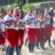 Girls softball team lined up on a dusty field, wearing white jerseys and red pants, smiling and clapping together in celebration.