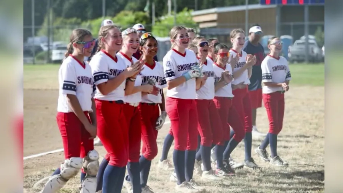 Girls softball team lined up on a dusty field, wearing white jerseys and red pants, smiling and clapping together in celebration.