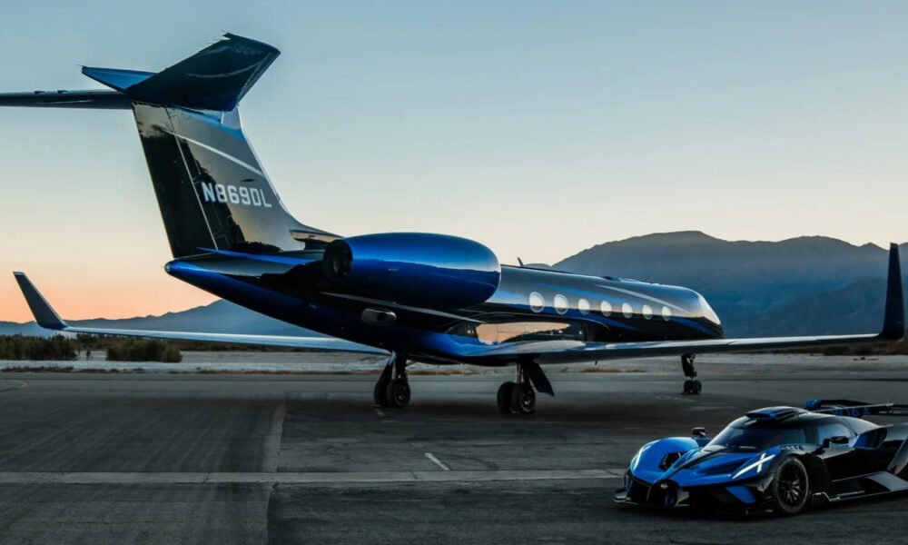 Private jet with a blue engine and a blue-tail design on a tarmac at sunset, with a sleek blue sports car parked nearby and mountains in the background