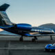 Private jet with a blue engine and a blue-tail design on a tarmac at sunset, with a sleek blue sports car parked nearby and mountains in the background