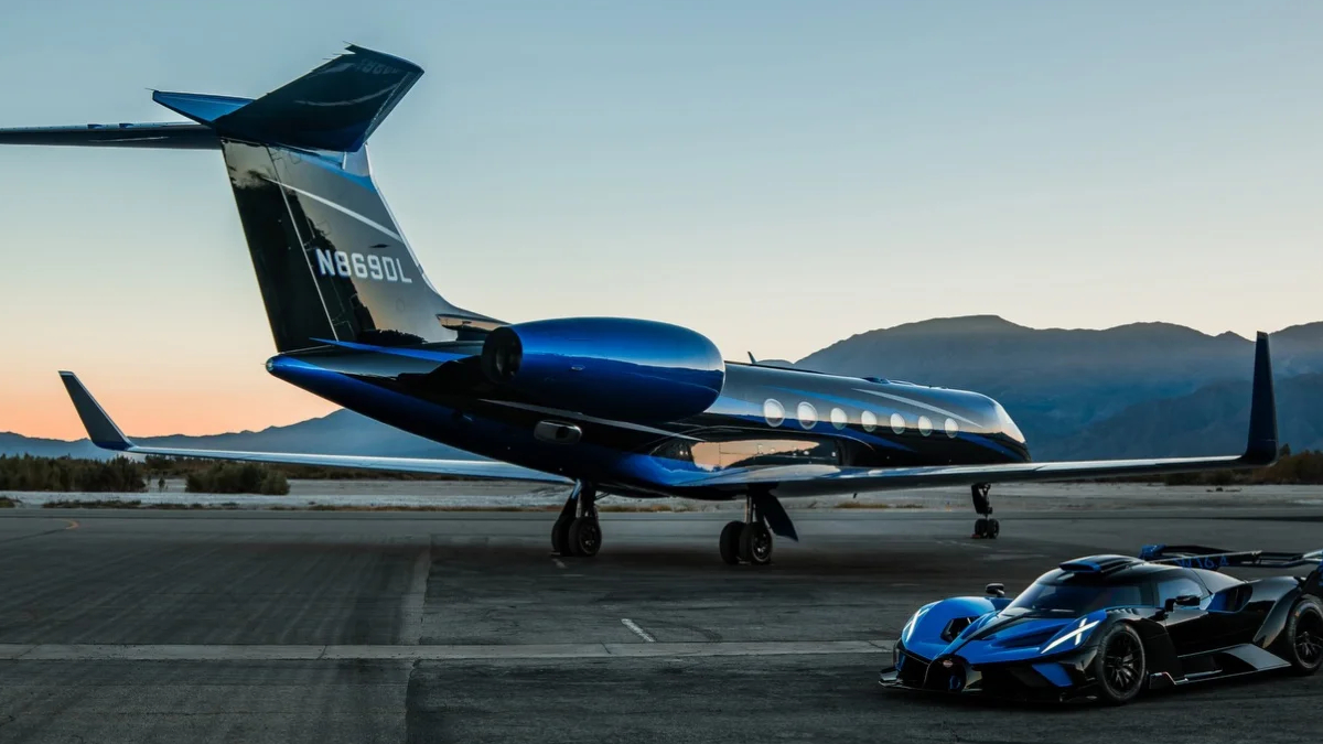 Private jet with a blue engine and a blue-tail design on a tarmac at sunset, with a sleek blue sports car parked nearby and mountains in the background