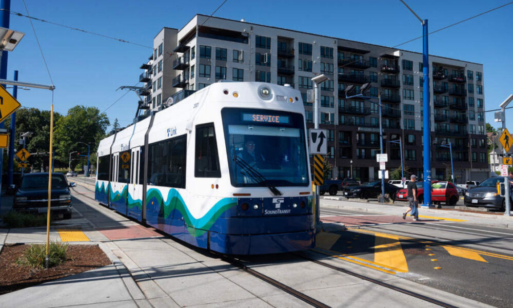White and blue modern tram on tracks at a city intersection, with apartment buildings in the background on a sunny day.