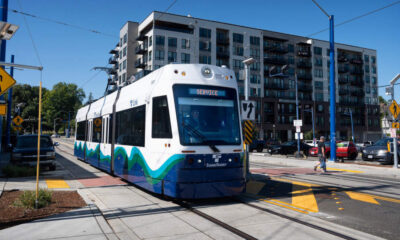 White and blue modern tram on tracks at a city intersection, with apartment buildings in the background on a sunny day.