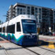 White and blue modern tram on tracks at a city intersection, with apartment buildings in the background on a sunny day.