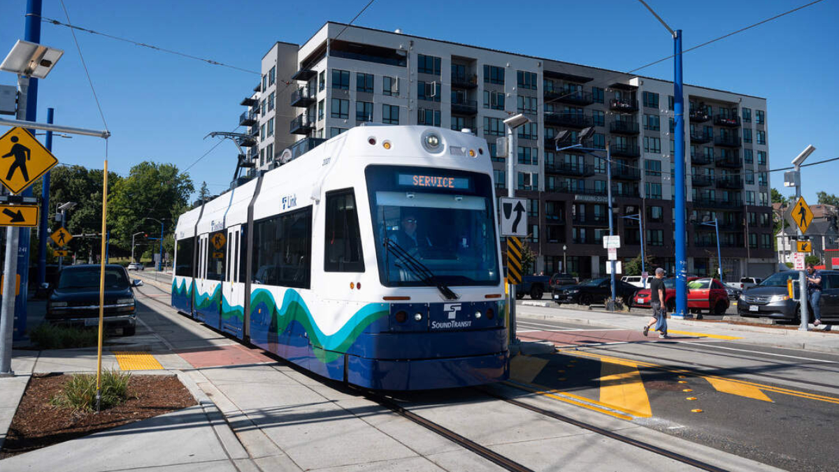 White and blue modern tram on tracks at a city intersection, with apartment buildings in the background on a sunny day.
