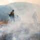 Firefighters in yellow gear using hoses to fight a smoky wildfire in a hilly landscape.