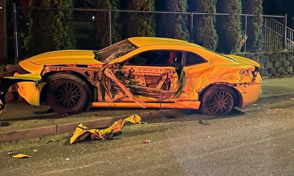 Yellow sports car with severe damage along the driver's side, exposed interior, and crumpled doors on a street at night; debris scattered nearby.