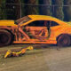 Yellow sports car with severe damage along the driver's side, exposed interior, and crumpled doors on a street at night; debris scattered nearby.