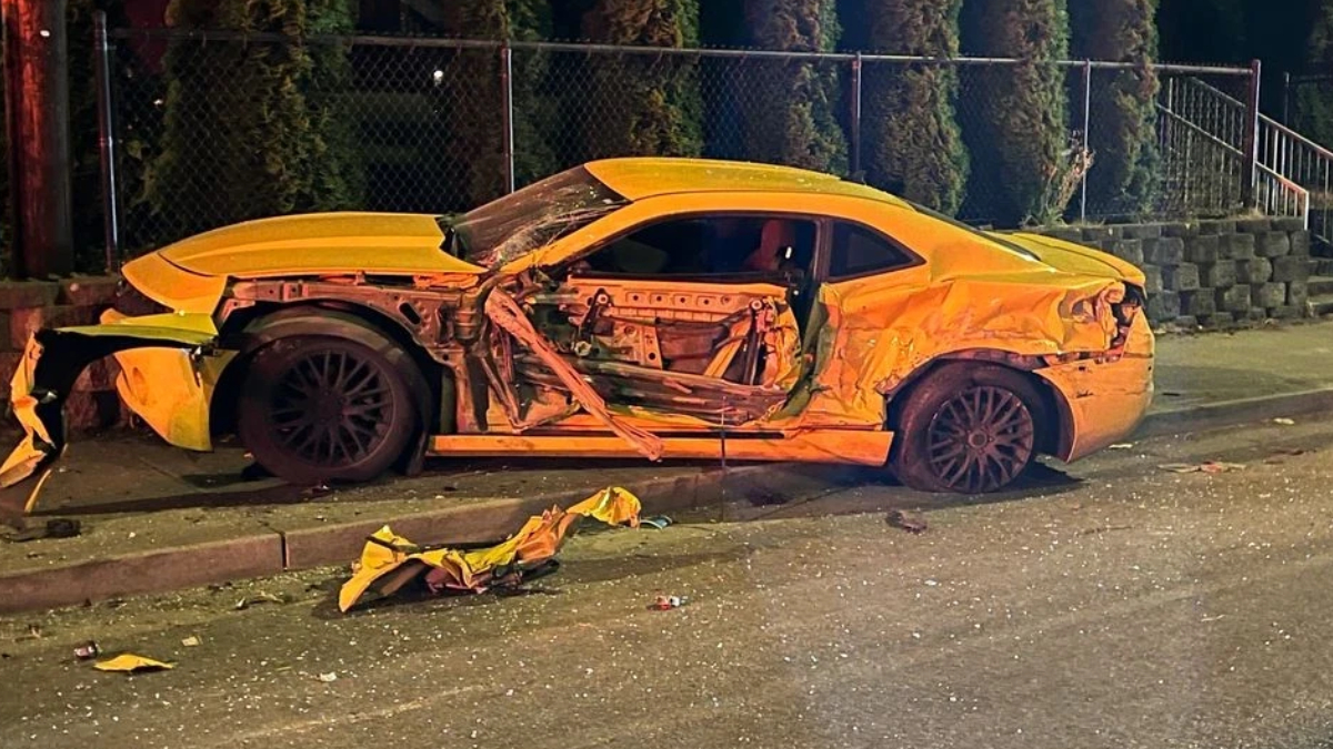 Yellow sports car with severe damage along the driver's side, exposed interior, and crumpled doors on a street at night; debris scattered nearby.