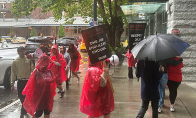 Group of protesters in red ponchos marching on a rainy city sidewalk, holding signs that say 'On Strike' and 'One Job Should Be Enough'.